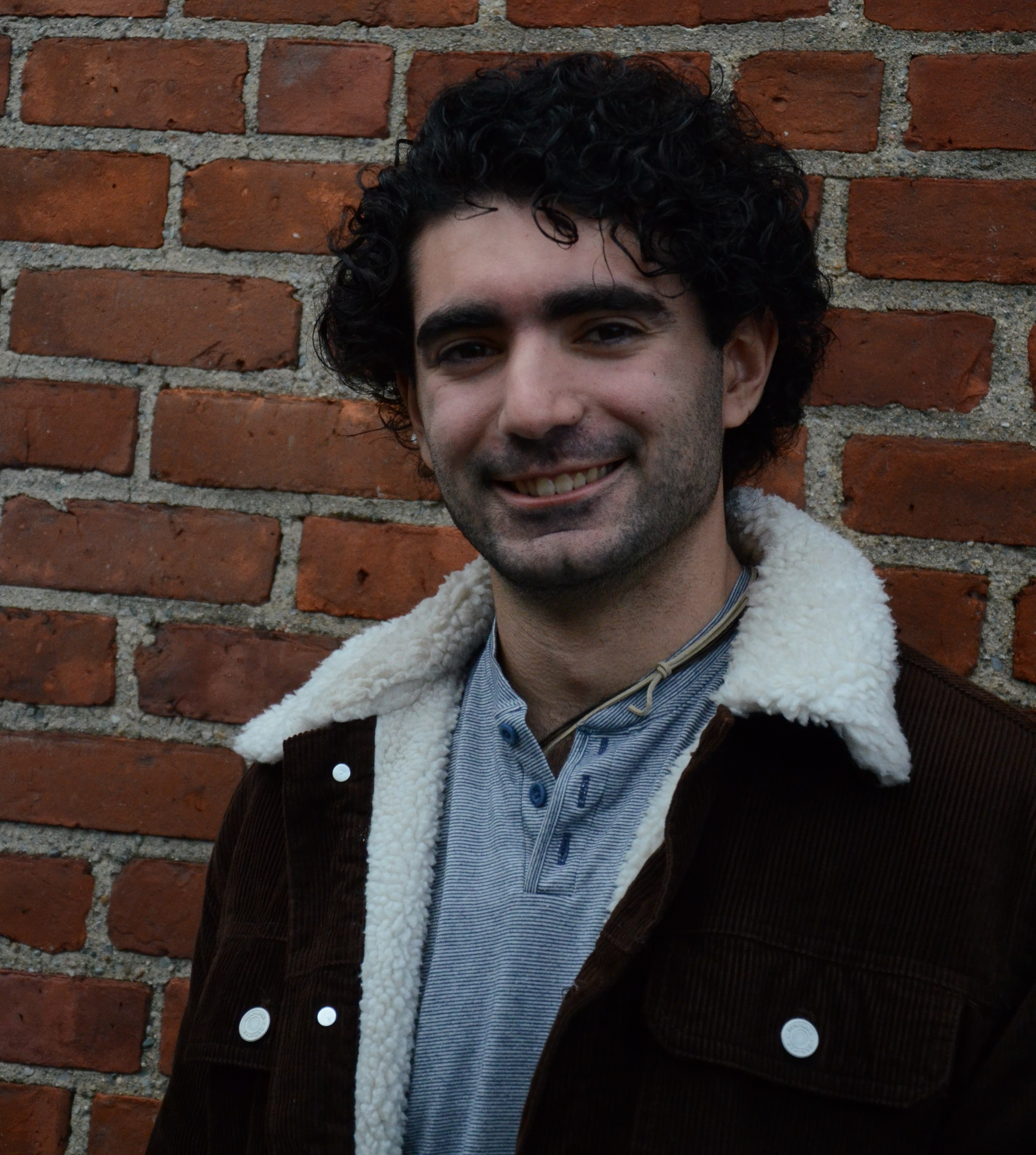 A professional photo of Matthew Blanco. He is smiling at the camera in front of a brick wall.